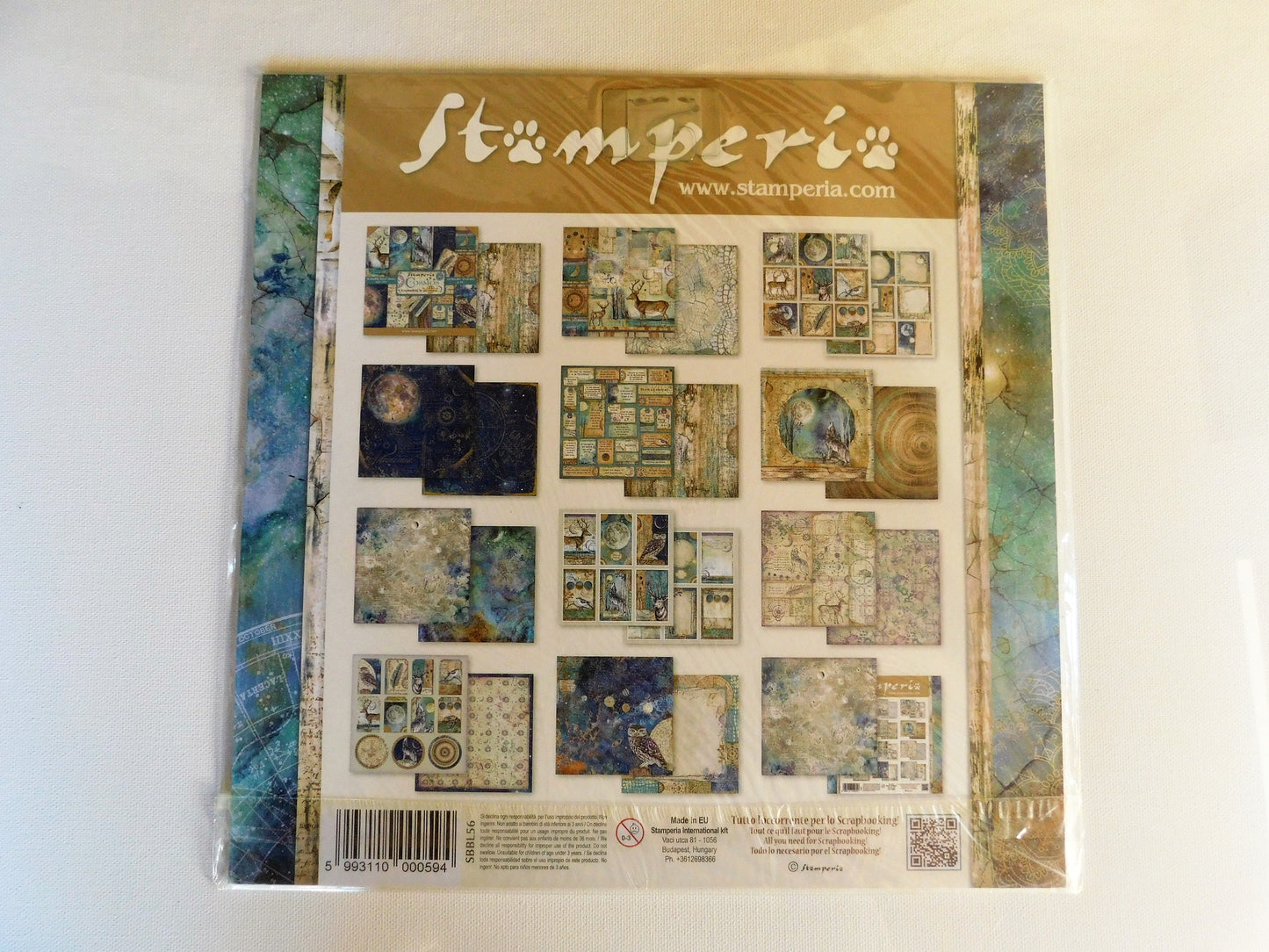 A packaged Stamperia scrapbooking paper sheet on a plain white background. The sheet features a collage of various textured paper designs in earthy tones of beige, blue, and green, framed by a decorative border.