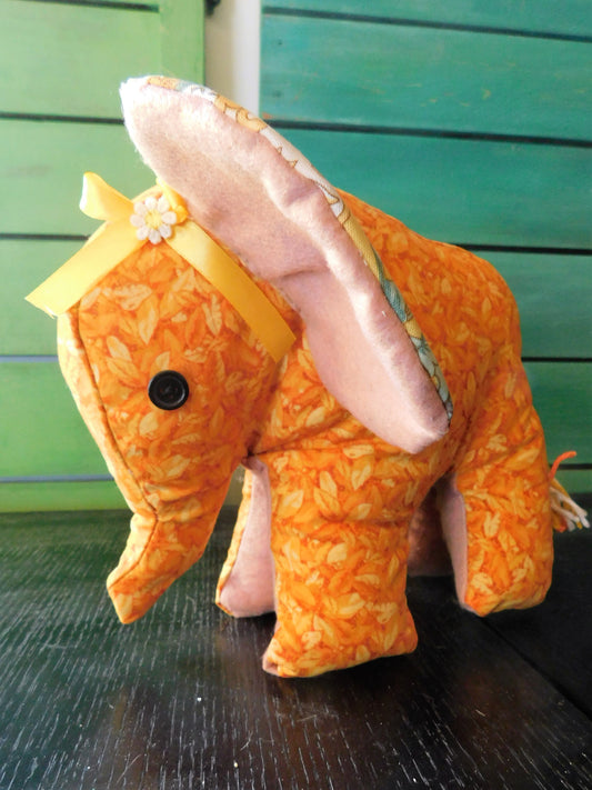 A handmade fabric elephant plush toy with an orange and yellow floral pattern, pink inner ears, and a yellow ribbon bow on its head, standing on a dark wooden surface against a green wooden background.
