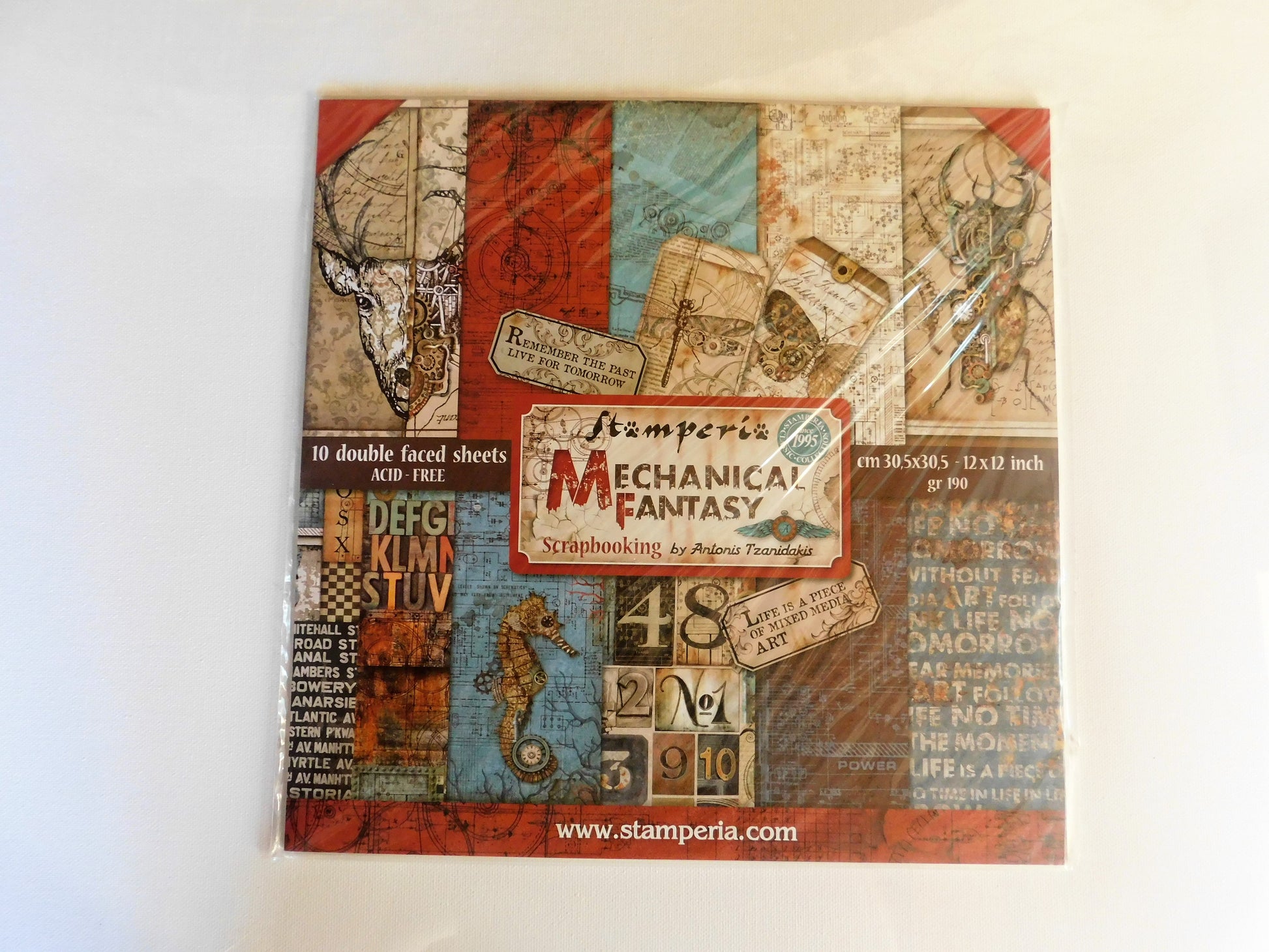 A square scrapbooking paper sheet set in plastic wrapping, featuring a vintage mechanical and industrial collage design with red, blue, beige, and brown tones, labeled 'Stamperia Mechanical Fantasy'.
