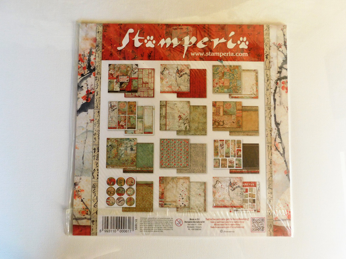 A packaged scrapbook paper set from Stamperia, features a red header with the white 'stamperia' logo and website, and displays a grid of various patterned paper designs in earthy tones of beige, green, and red.