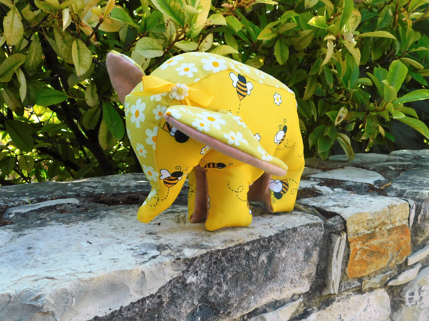A small yellow fabric elephant toy with white daisy and bee prints, pink ears, and a matching yellow bow, standing on a stone wall with green foliage in the background.