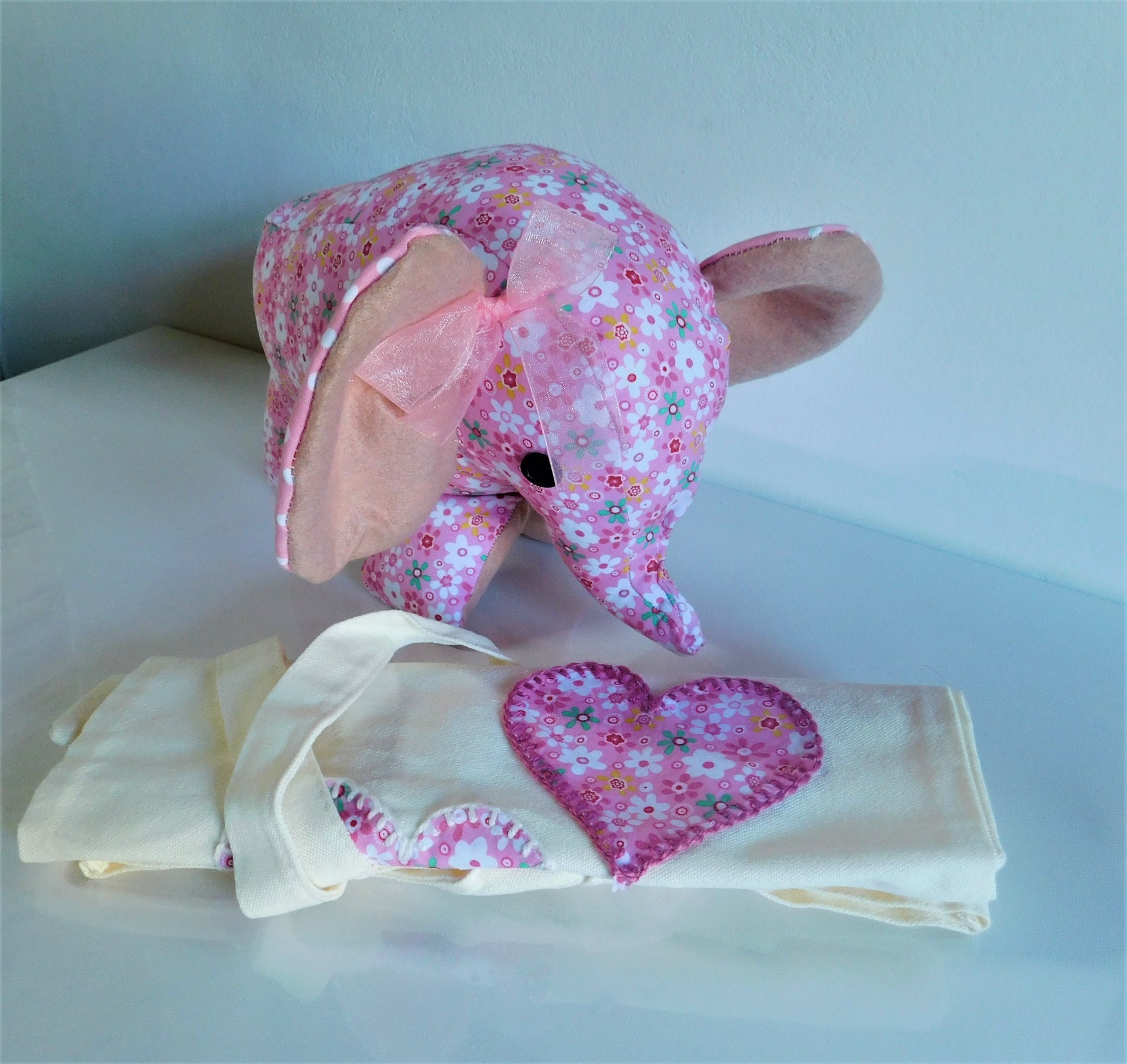 A pink floral elephant plush toy with a matching pink bow, posed above folded cream-colored baby blankets featuring a matching pink floral heart applique.