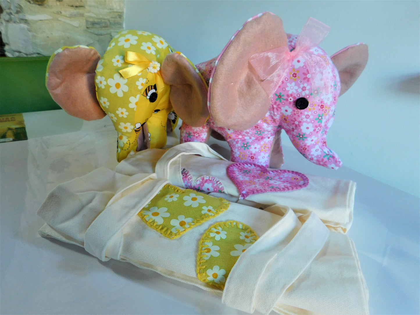 A baby elephant-themed fabric set featuring a plush elephant toy with a yellow floral pattern and pink ears, a matching pink floral plush elephant, and cream-colored fabric accessories with white floral patches.