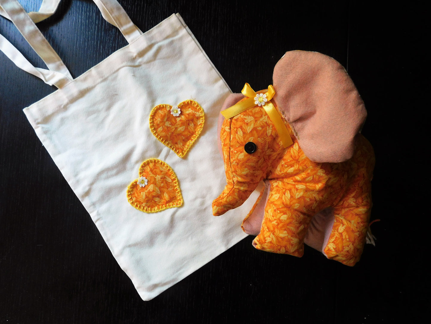 A white canvas tote bag with two orange and yellow patterned heart patches, shown next to a matching orange and yellow patterned elephant plush toy on a dark background.