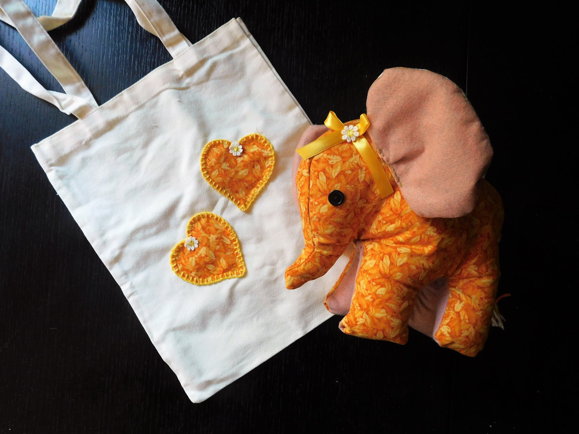A white canvas tote bag with two orange and yellow patterned heart patches, shown next to a matching orange and yellow patterned elephant plush toy on a dark background.